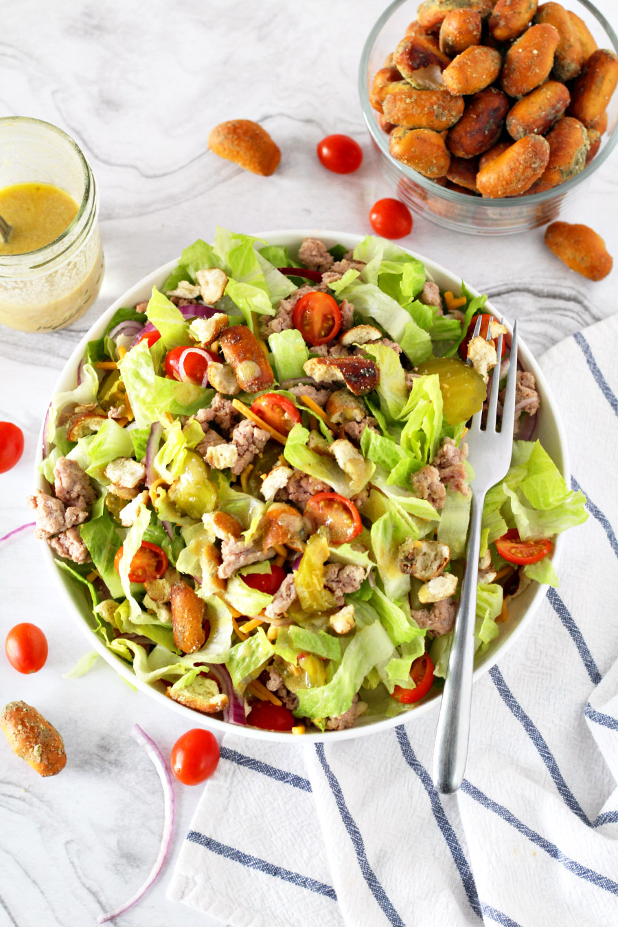 Cheeseburger Salad with Pretzel Croutons in a bowl next to dressing and a bowl of Garlic OMG! Pretzels.
