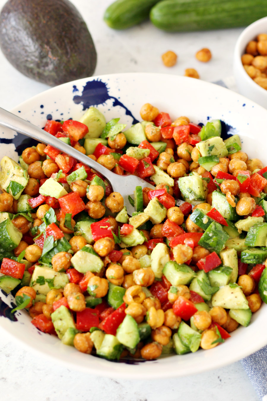 Fork siting in a bowl of Crispy Chickpea and Avocado Salad. Avocado, cucumbers, and crispy chickpeas in the background.