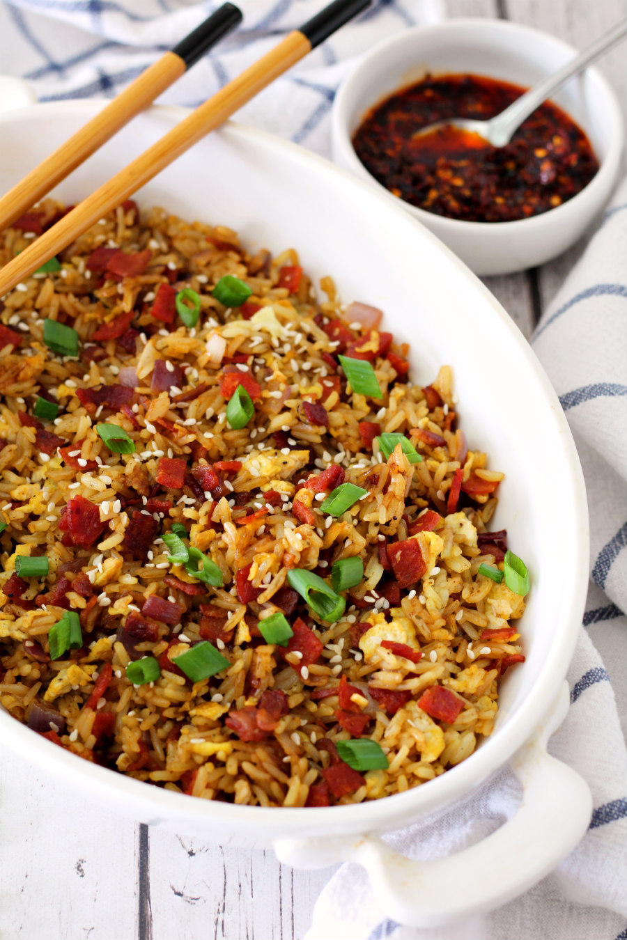Bacon and Chorizo Fried Rice in a white serving dish with chopsticks sitting on the edge. Hot chili crisp and kitchen towels in the background.