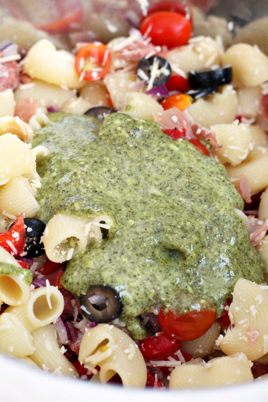 Chiocciole pasta in a mixing bowl with red bell pepper, onion, olives, grape tomatoes, and prosciutto with shredded Parmesan cheese and basil pesto.