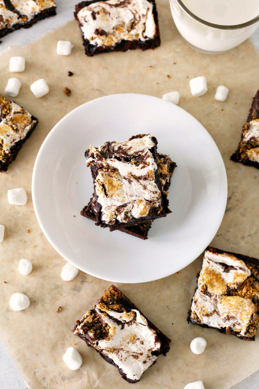 Flat lay photo of Easy S'mores Swirled Brownies stacked on a plate and scattered around on parchment paper with mini marshmallows.