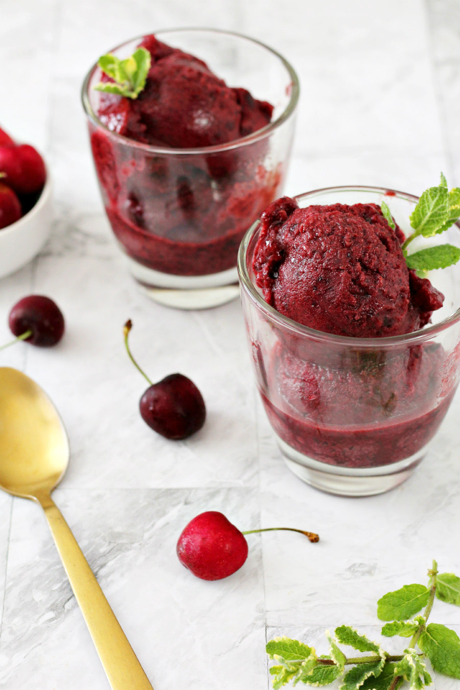 Easy Cherry Bourbon Sorbet served in glasses. Small bowl of fresh cherries, gold spoon, and fresh mint sit around the glasses.