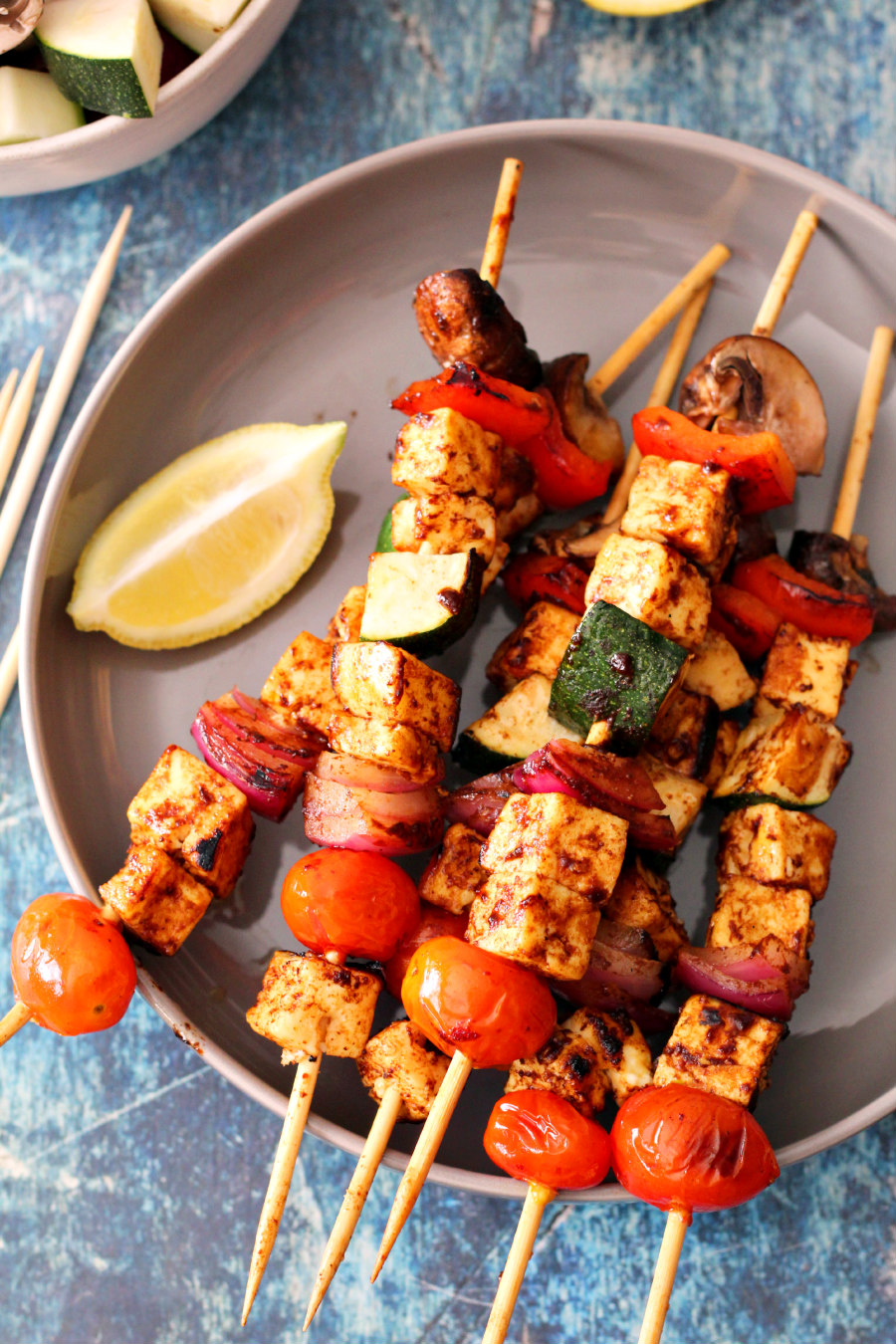 Flat lay photo of Grilled Paneer Veggie Kebabs on a plate. Bamboo skewers, bowl of prepared veggies, and lemon slices surround the plate.