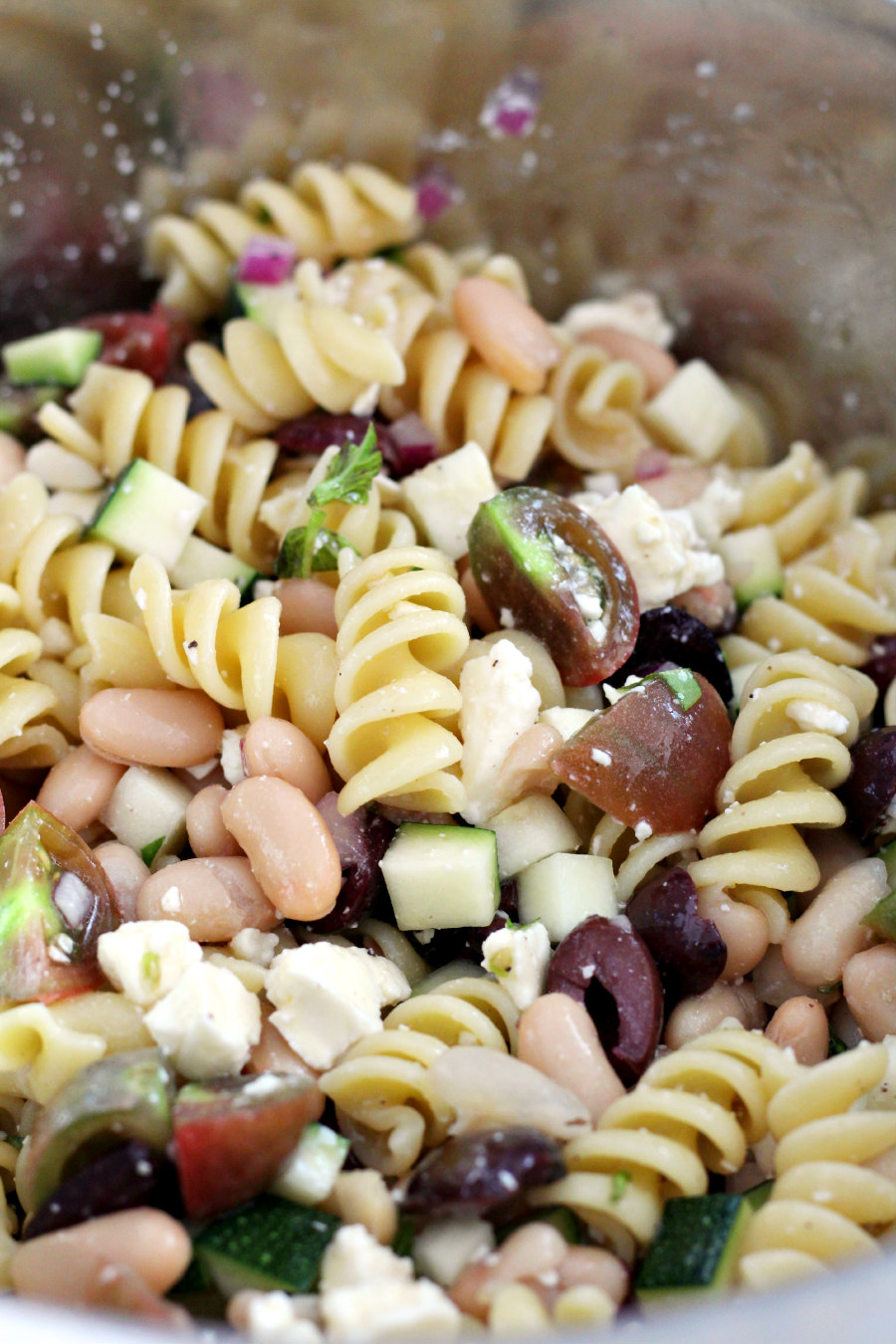 Rotini pasta, cannellini beans, red onion, basil, zucchini, cherry tomatoes, and kalamata olives, and crumbled feta cheese in a metal mixing bowl.