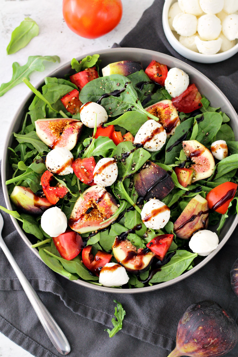 Flat lay photo of Trader Joe's Inspired Black Fig and Balsamic Salad pictured with fork, arugula leaves, tomato, fresh mozzarella balls, and a fresh fig.