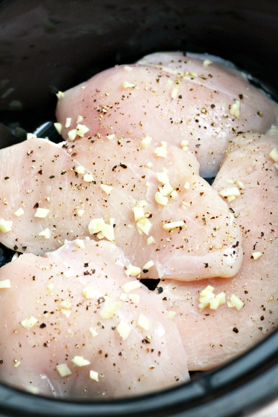 Raw chicken breasts in a slow cooker seasoned with salt and pepper and sprinkled with fresh chopped garlic.