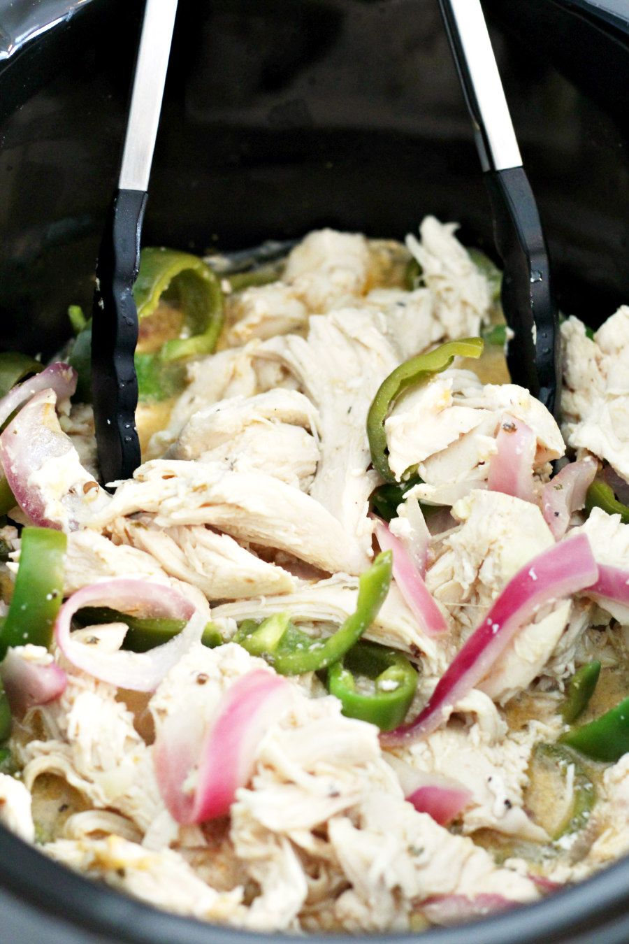 Chicken shredded in slow cooker with peppers and onions. Tongs sit inside slow cooker.