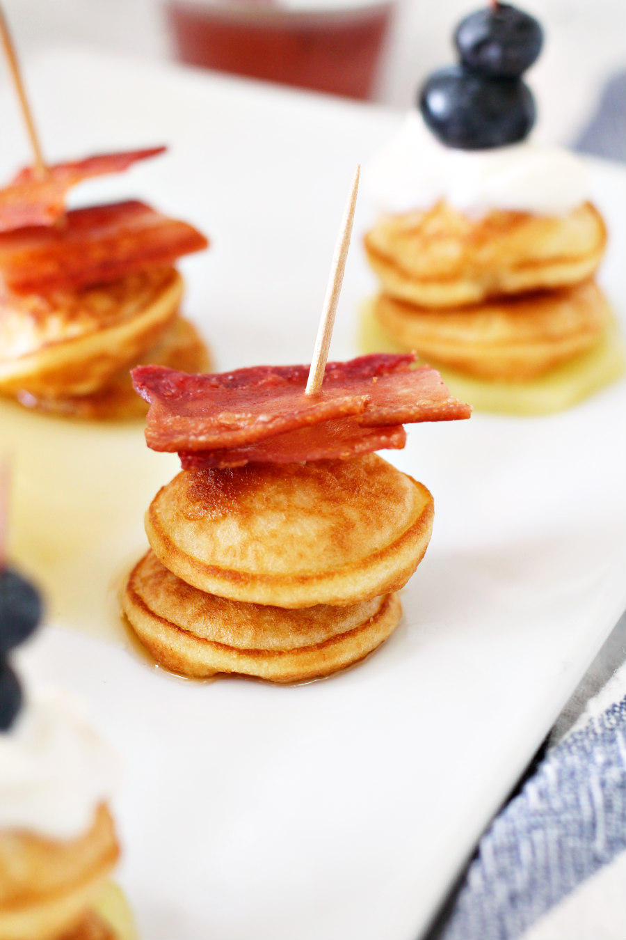 Close up of mini pancake stack topped with maple syrup and bacon. Other pancake stacks are arranged on a serving dish.