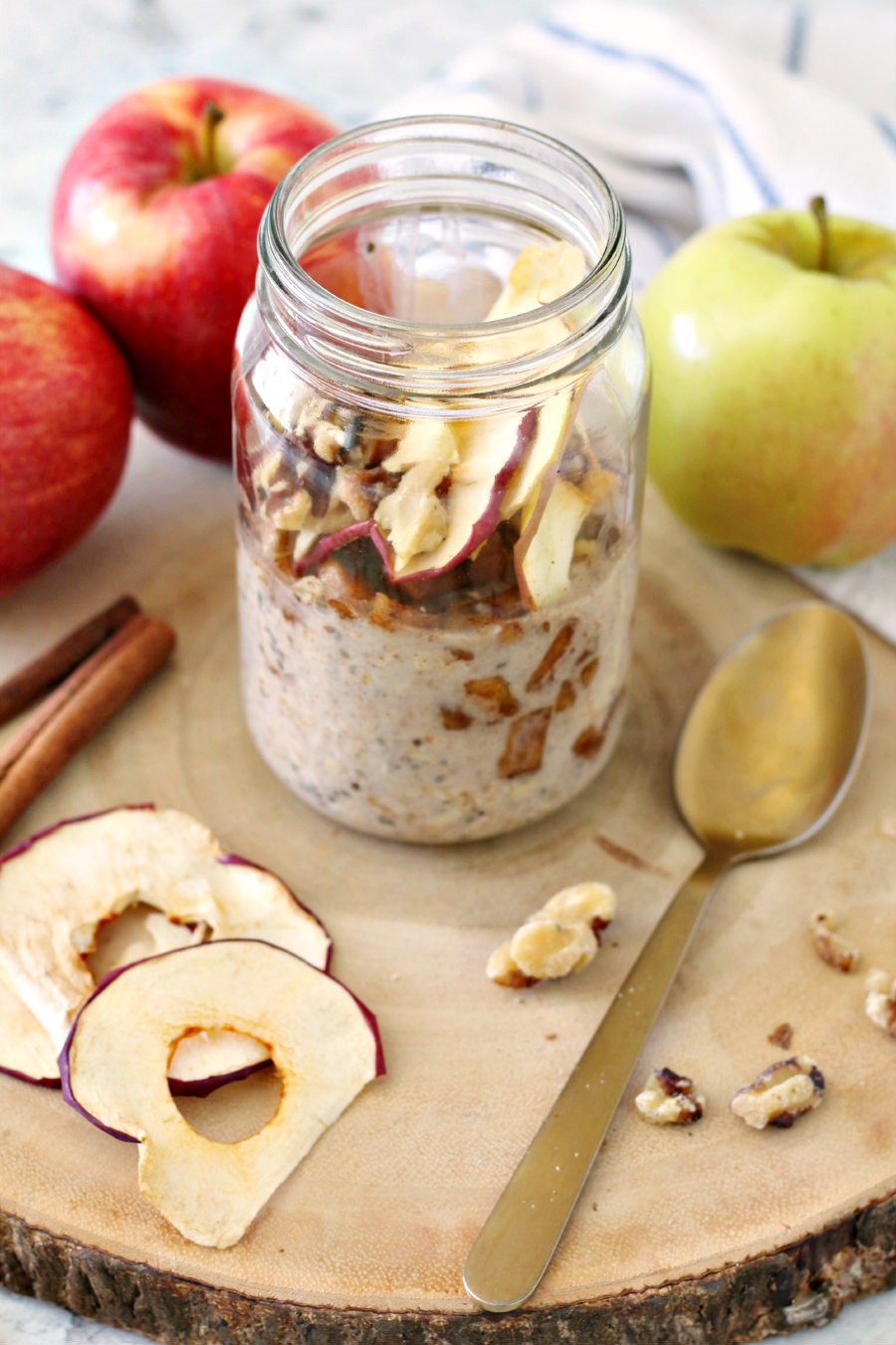 Apple Pie Overnight Oats on a wood slice with apples, dried apples, cinnamon sticks, walnuts, and a gold spoon in photo.