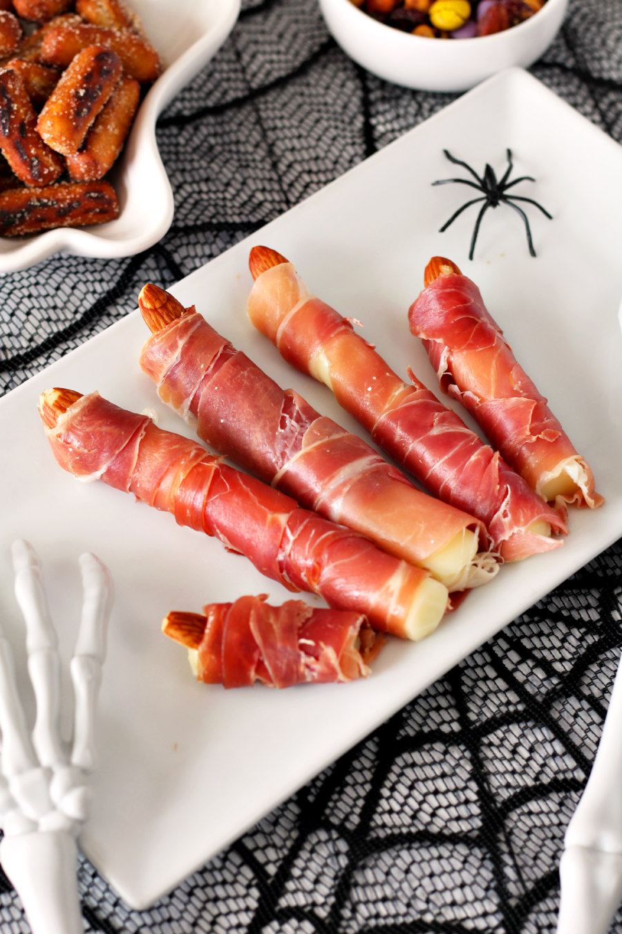 Prosciutto Wrapped Mozzarella Fingers on a serrving plate with plastic spiders, skeleton hands, and small bowls of snacks in background.