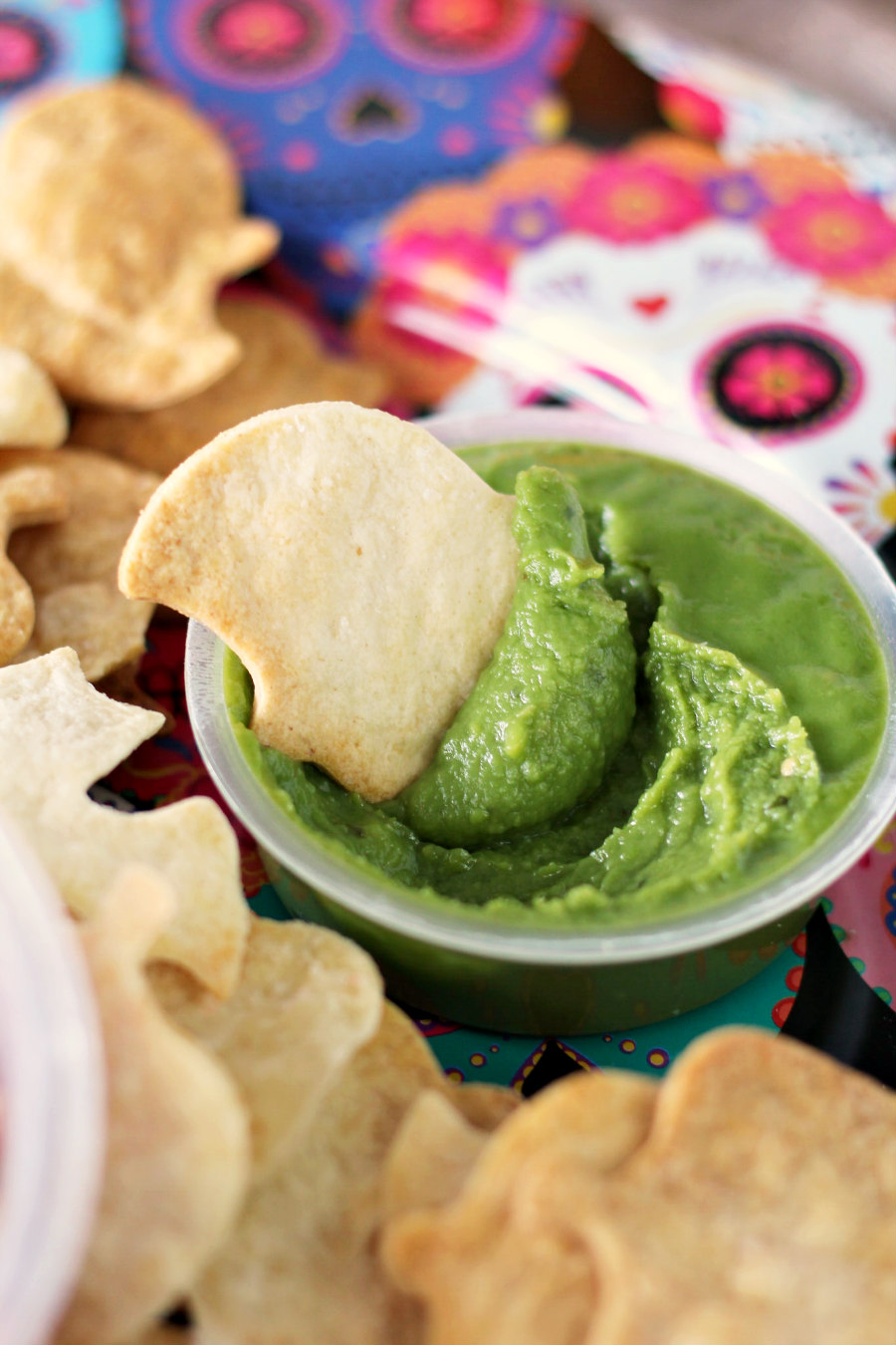 Air Fryer Halloween Tortilla Chip dipped into cup of guacamole. Chips in background on sugar skull plate.