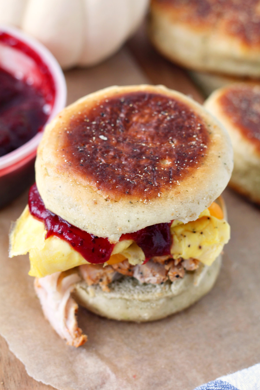 Thanksgiving Leftovers Breakfast Sandwich on parchment lined wooden board. Container of cranberry sauce, white pumpkin, and Stuffin' Muffins also in photo.