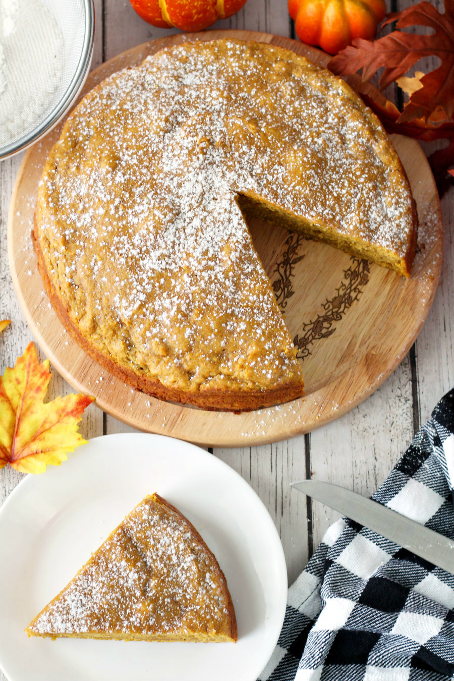 Pumpkin Spice Ricotta Cake on wooden board. Slice of cake pictured on small plate. Checkered towel, knife, powdered sugar, and autumn leaves also in photo.