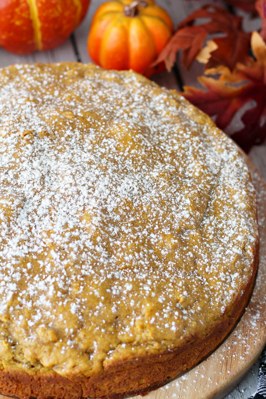 Close up of uncut Pumpkin Spice Ricotta Cake on wooden board. Autumn leaves and pumpkins in background.